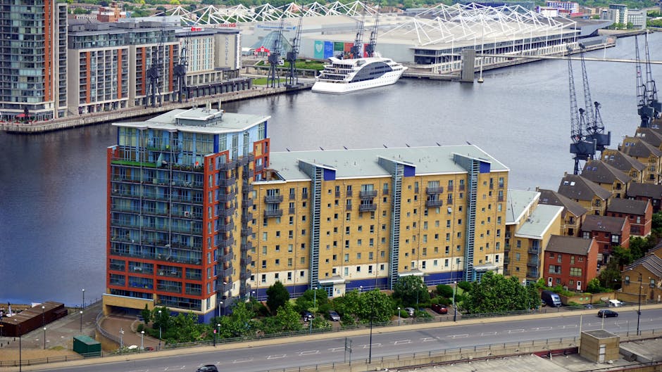 A view of a modern city skyline in Docklands with high-rise buildings made of glass and steel, reflecting off the calm water of a river or marina. The buildings vary in height and design, with some fe