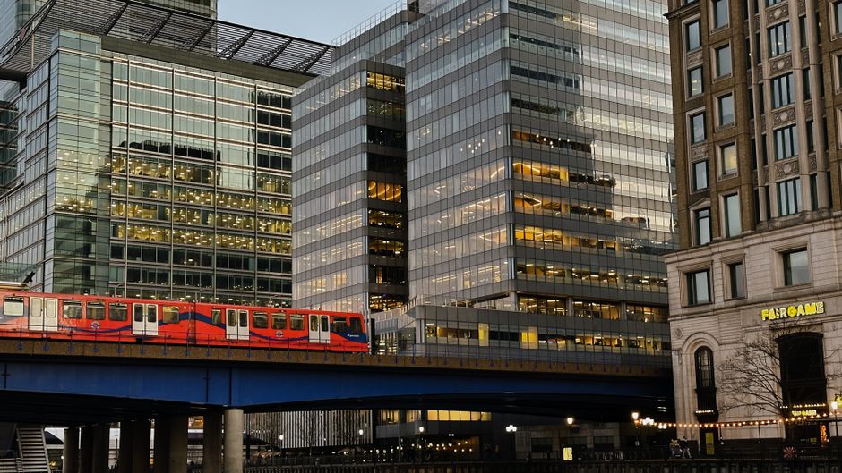 A view of modern office skyscrapers with glass facades reflecting city lights in Docklands, London, taken during dusk. In the foreground, a red London train travels along a blue elevated railway track. To the right, an older stone building labeled 'FARBOAM' stands adjacent to contemporary glass structures, creating a contrast between historic and modern architecture. The scene is well-lit with ambient lighting from the buildings and streetlights, highlighting the clean surfaces of the glass and steel materials, with the train and surrounding area appearing tidy and well-maintained. This urban landscape exemplifies a typical commercial environment in Docklands, with a focus on the sleek, reflective surfaces of the office buildings and the organized, safe railway infrastructure, illustrating cleanliness and modernity in cityscape design, as seen through the lens of a professional cleaning service like Cleaner Docklands.