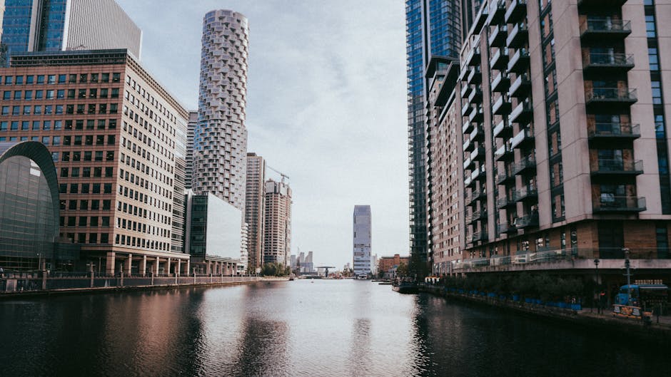 A daytime view of the Canary Wharf waterfront in Docklands, featuring modern high-rise office buildings constructed from materials such as glass, steel, and concrete. The scene includes reflections of the buildings on the calm water, with reflections of glass facades and metallic surfaces visible on the surface. The skyline is illuminated by natural light, highlighting the cleanliness and sleekness of the urban environment. To the right, a tall building with a mix of glass and structural materials features balconies, while on the left, a building with a grid-like window pattern and rounded architectural elements can be seen. The overall scene emphasizes the commercial architectural landscape of Docklands, suitable for visual references in deep cleaning and surface sanitisation related to office environments, as promoted by Cleaner Docklands.
