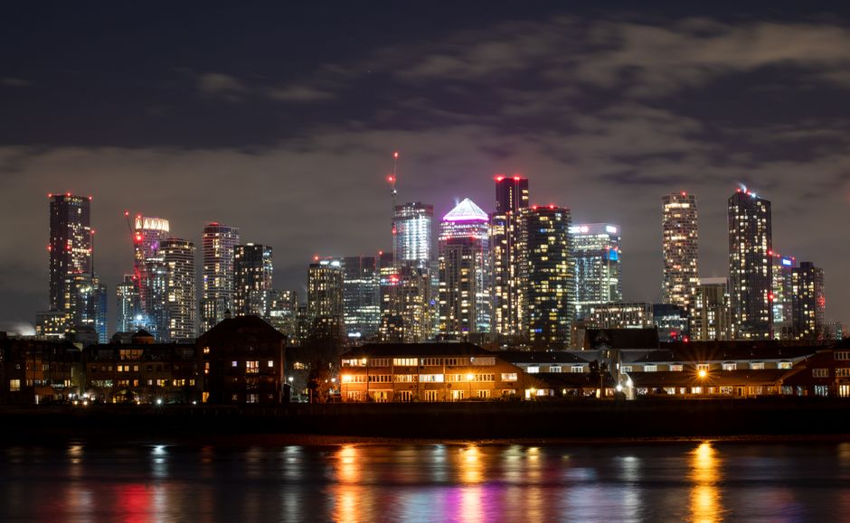 A view of modern office skyscrapers with glass facades reflecting city lights in Docklands, London, taken during dusk. In the foreground, a red London train travels along a blue elevated railway track. To the right, an older stone building labeled 'FARBOAM' stands adjacent to contemporary glass structures, creating a contrast between historic and modern architecture. The scene is well-lit with ambient lighting from the buildings and streetlights, highlighting the clean surfaces of the glass and steel materials, with the train and surrounding area appearing tidy and well-maintained. This urban landscape exemplifies a typical commercial environment in Docklands, with a focus on the sleek, reflective surfaces of the office buildings and the organized, safe railway infrastructure, illustrating cleanliness and modernity in cityscape design, as seen through the lens of a professional cleaning service like Cleaner Docklands.