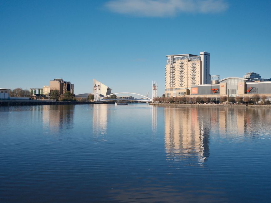 A panoramic view of the Docklands area on Westferry Road, Docklands, featuring modern multi-story buildings, including a prominent high-rise with numerous balconies, a shopping mall with store signs, 