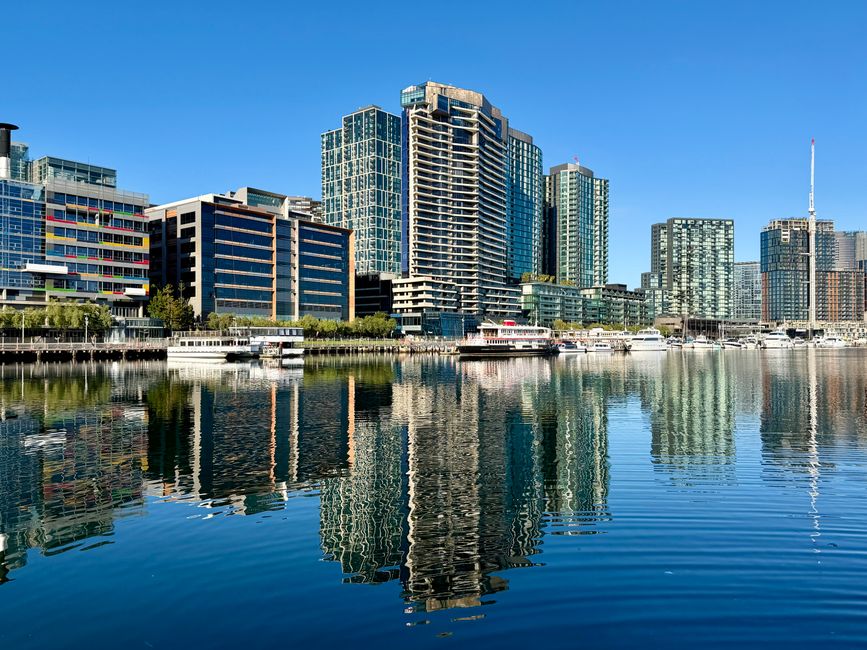 A panoramic view of the Docklands skyline featuring modern glass skyscrapers with reflective surfaces, some with unusual architectural shapes. The calm water in the foreground mirrors the buildings an
